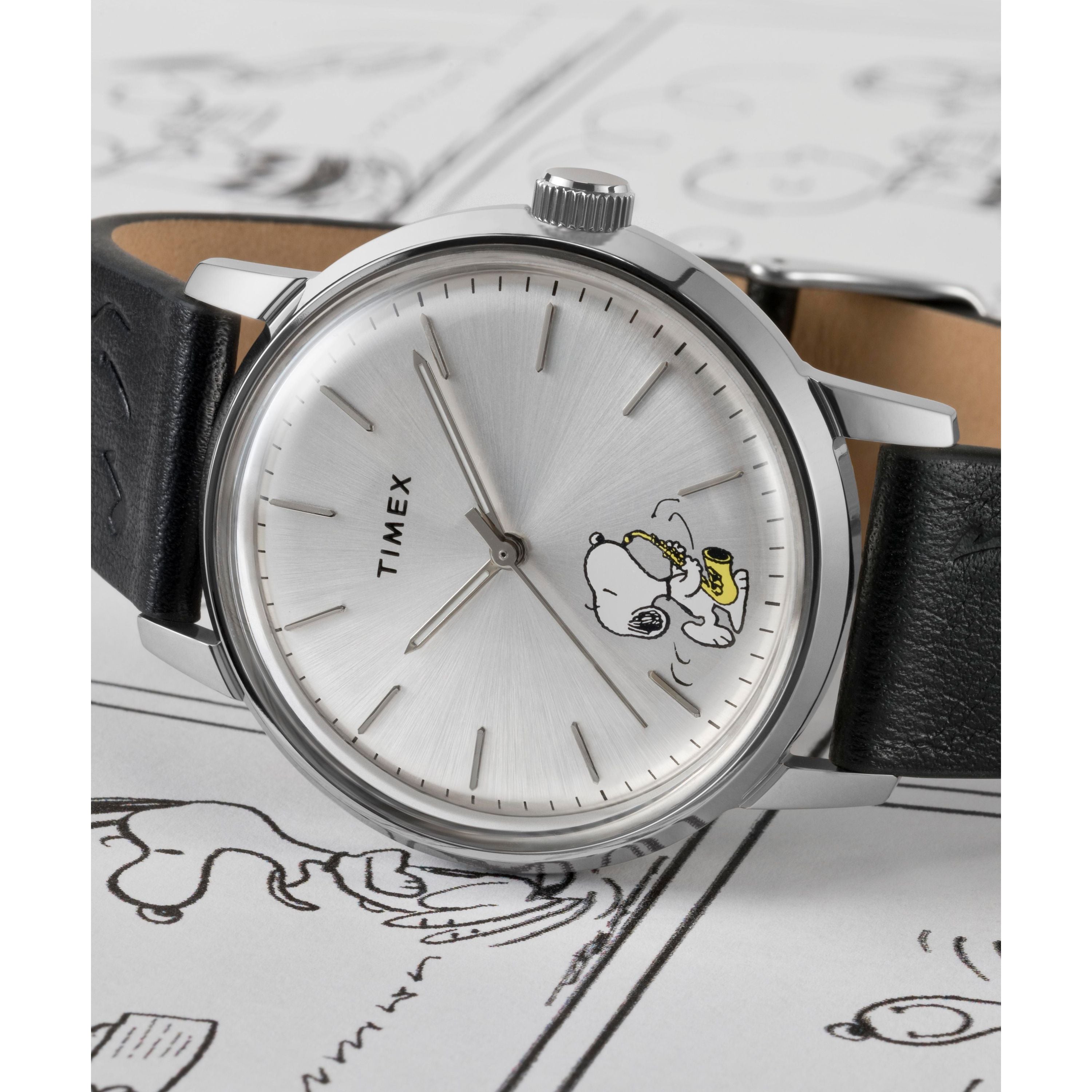 A Timex x Peanuts Marlin® Automatic Saxophonist watch by Timex features a silver dial, black leather strap, and Snoopy as a saxophonist on the face, displayed atop a surface decorated with Snoopy comic strip illustrations.