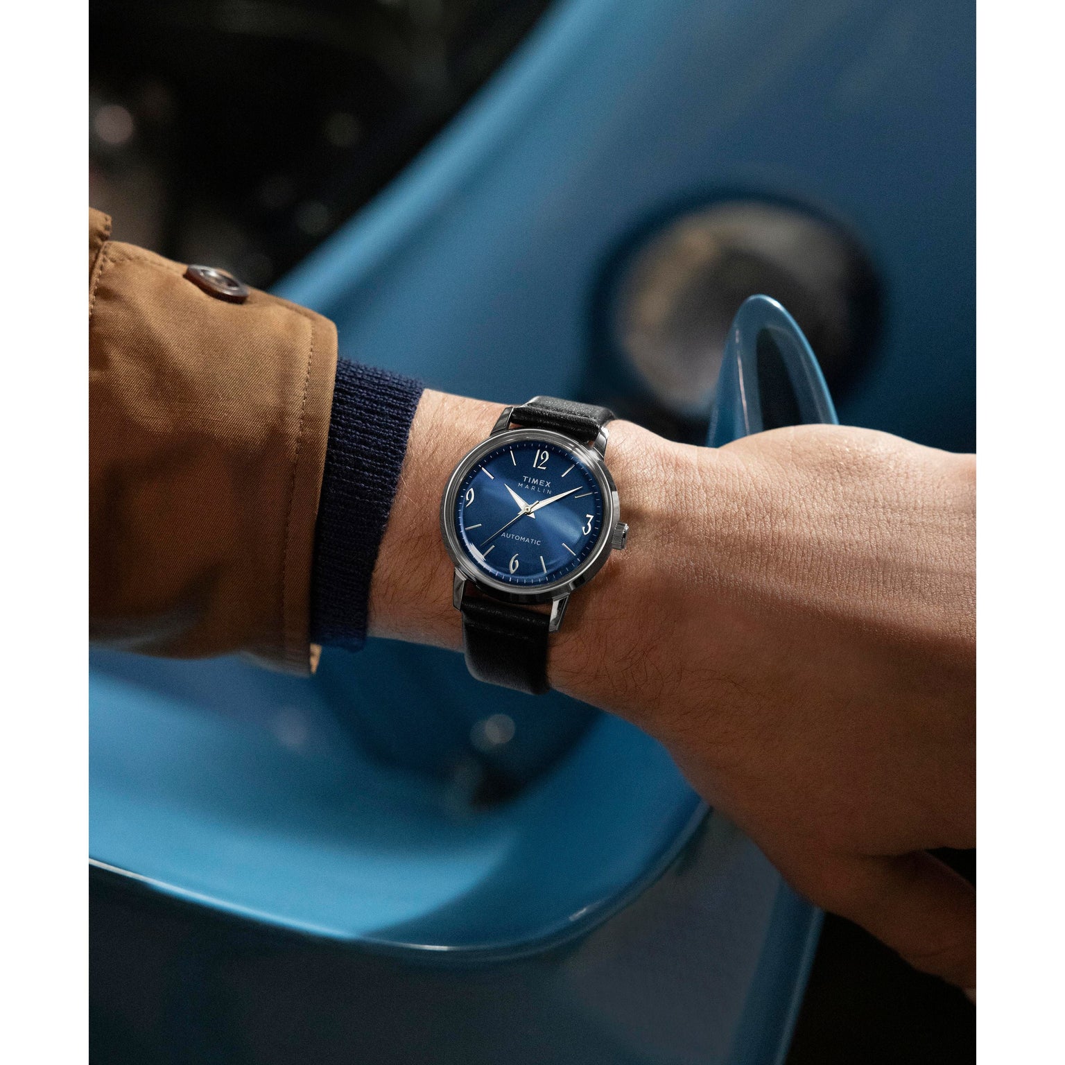 A person in a brown coat and navy sweater showcases the Timex Marlin® Automatic wristwatch, featuring a blue face and eco-friendly leather strap; a curved blue car with visible headlight is seen in the background.