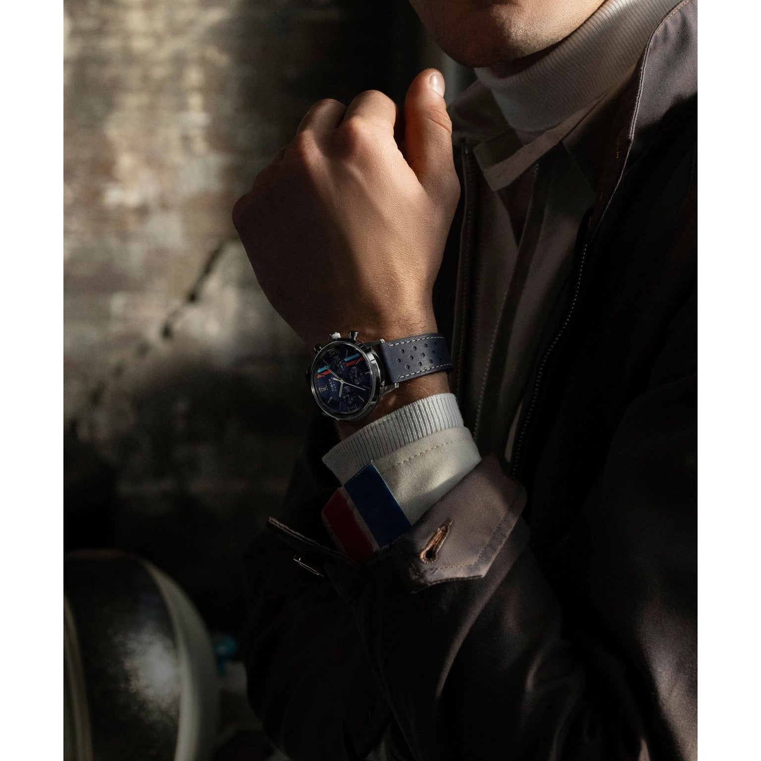 A person in a brown jacket and white turtleneck showcases the Timex Marlin® Chronograph Tachymeter with a perforated strap. Lighting highlights the watch and the French flag cuff detail against a dark, textured background.