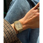 A person wearing a Timex Automatic 1983 E Line Reissue gold-tone dress watch sits with crossed legs, blue jeans, and a brown houndstooth coat. Sunlight highlights the watch’s Japanese automatic movement and their ring-adorned hand.