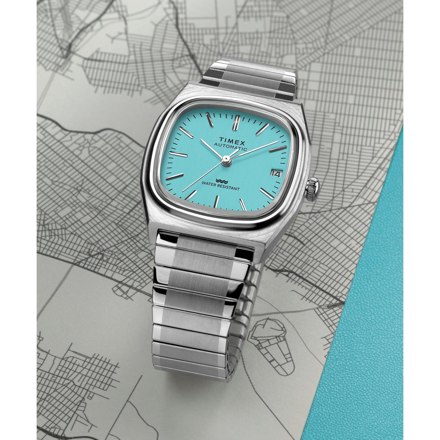 The Timex Automatic 1983 E Line dress watch, featuring a silver case, turquoise face, and metal band, is displayed atop a grayscale city map with a textured turquoise area on the right.