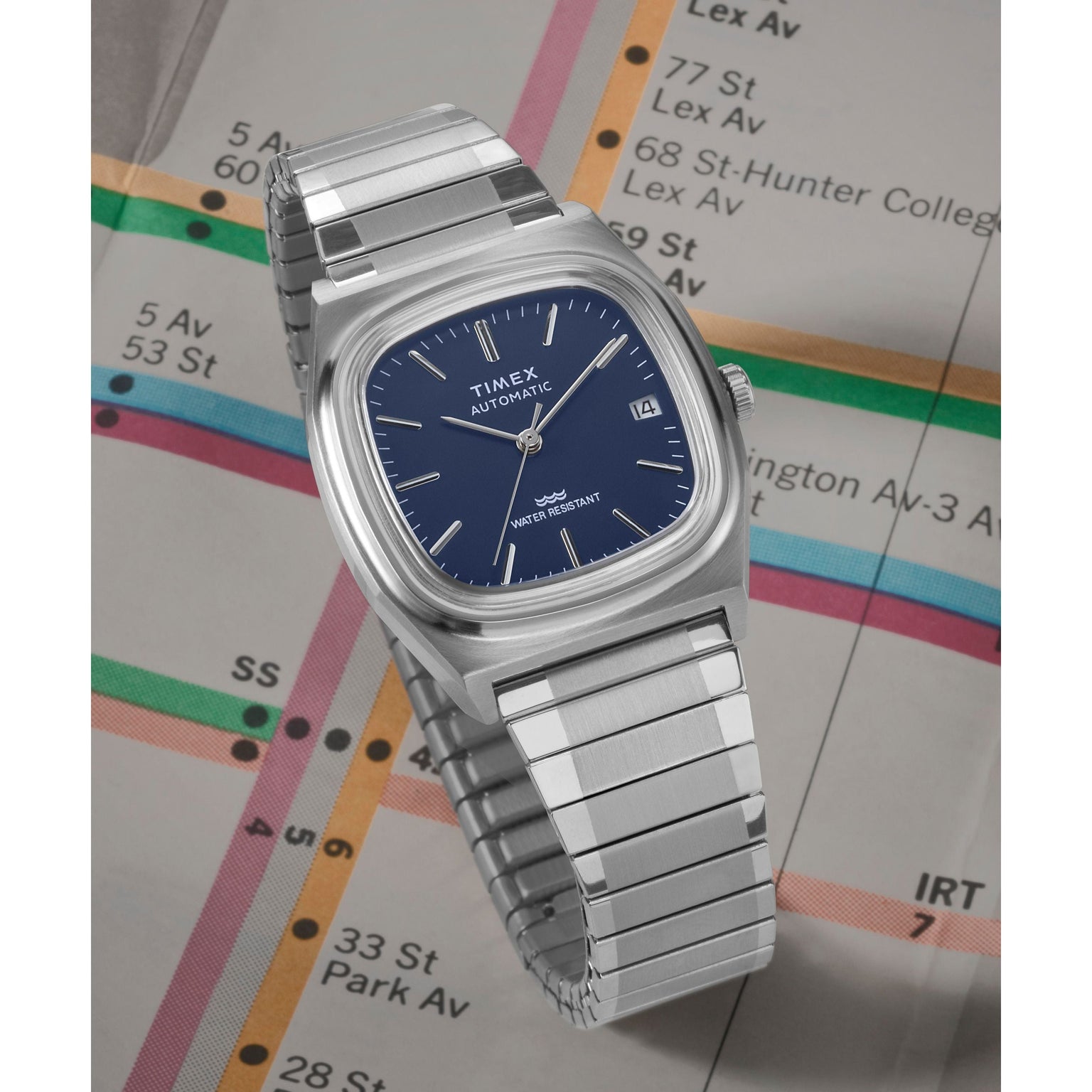 A Timex Automatic 1983 E Line wristwatch with a blue dial and date display, featuring a stainless-steel expansion band and square face, is placed on a New York City subway map.