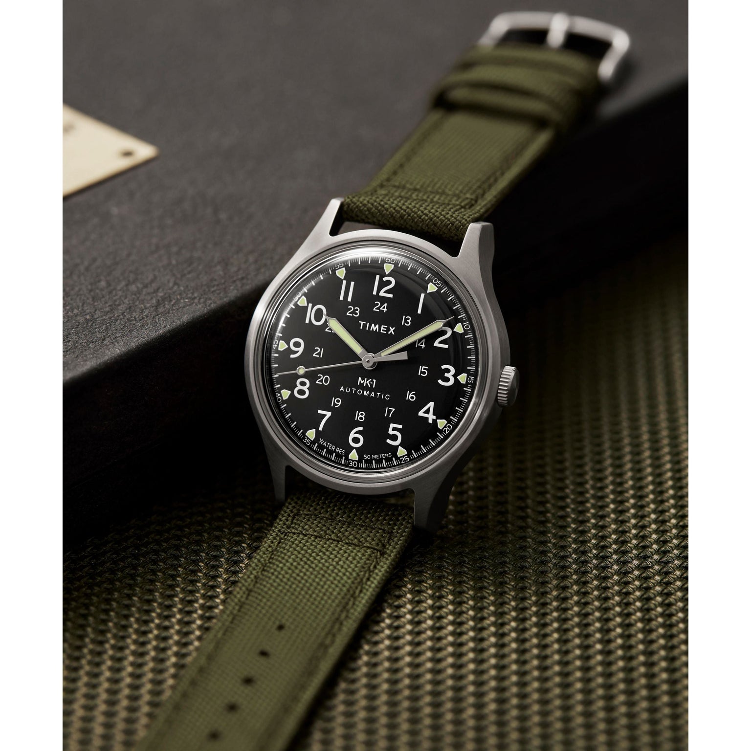 The Timex G SS AU SW MK1 40 BLK DL GRN STP 25, featuring a black dial, white numerals, olive green fabric strap, and stainless steel case, is shown on a textured green surface beside a black box.