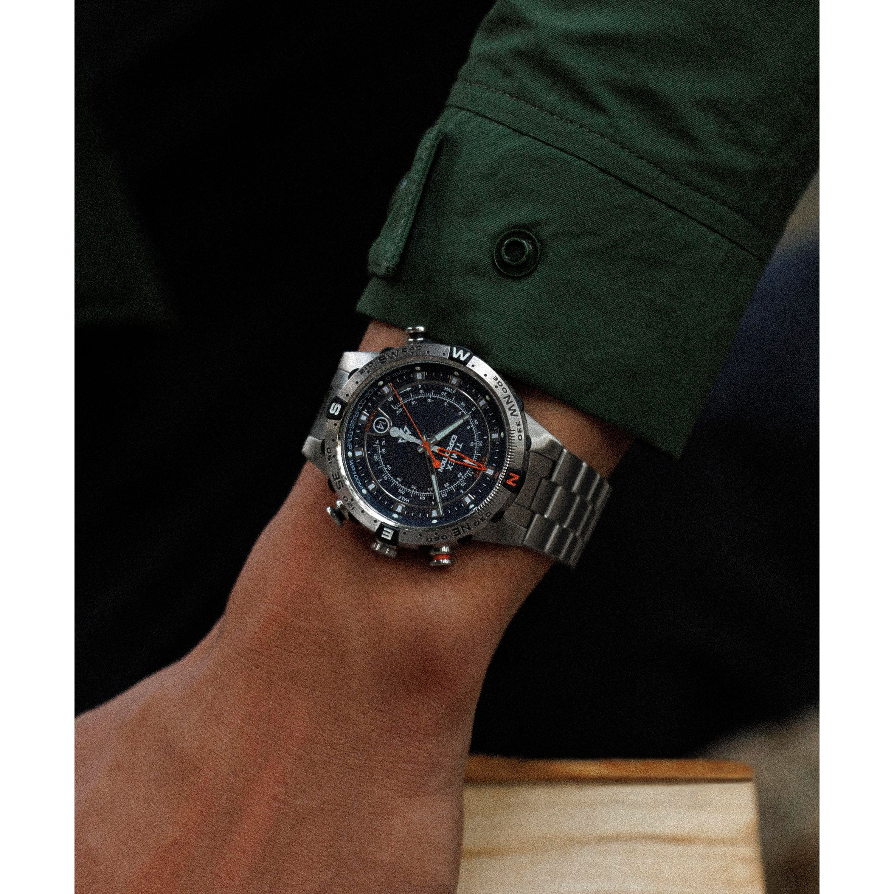 A close-up shows a wrist wearing the Timex Expedition Tide-Temp-Compass, featuring a stainless-steel case and dark dial with multiple markings. The wearer sports a dark green long-sleeve shirt, keeping focus on this elegant timepiece.