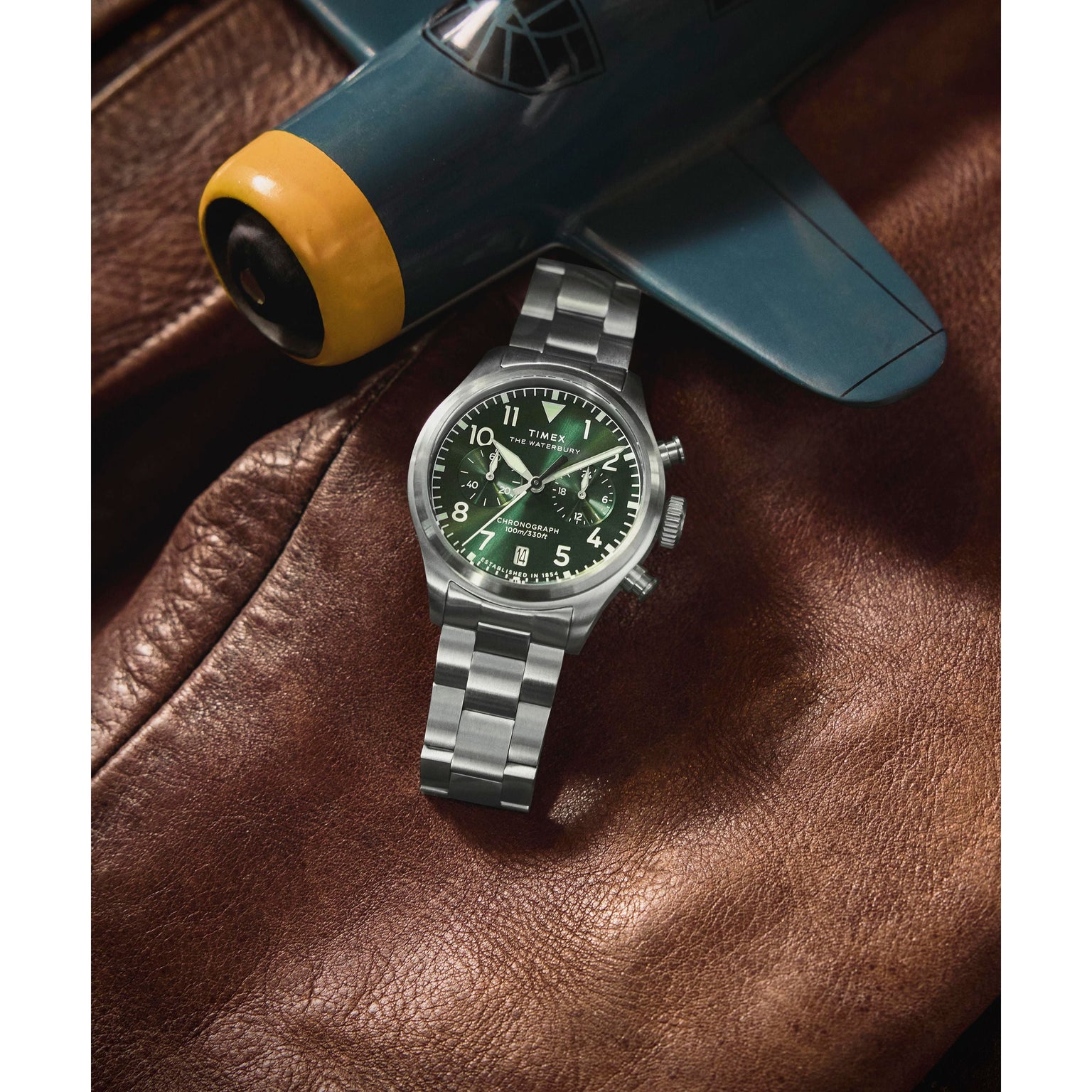 The Timex G SS QZ WATRBUY TRAD ACE CH 41 GRN DL 25, a stainless steel pilot's watch with a green dial, sits on brown leather beside a model airplane with a blue body and yellow nose.