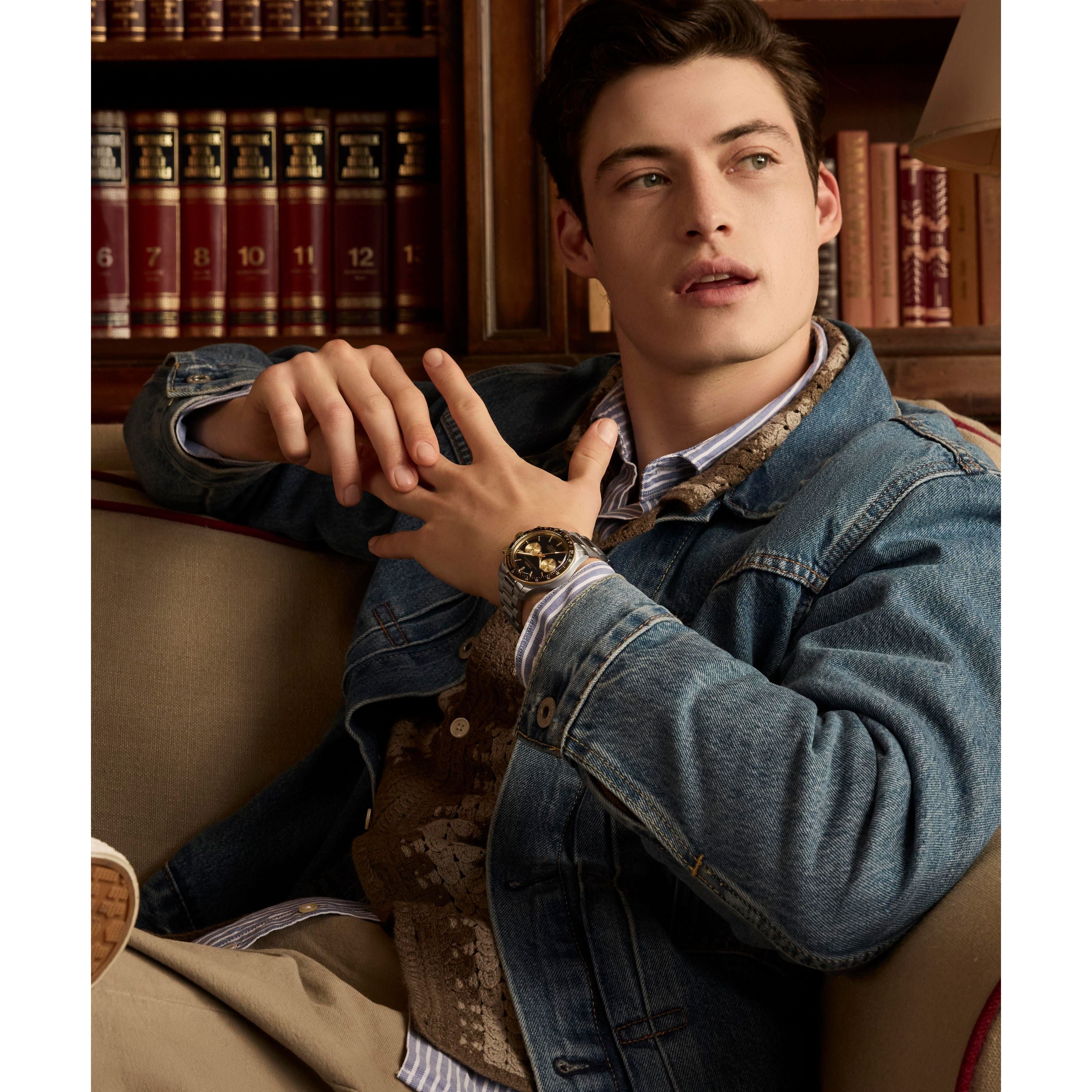 A young man in a denim jacket and a Timex Waterbury Heritage Chronograph sits on a sofa, gazing sideways. Behind him, shelves lined with books evoke a cozy library-like atmosphere.