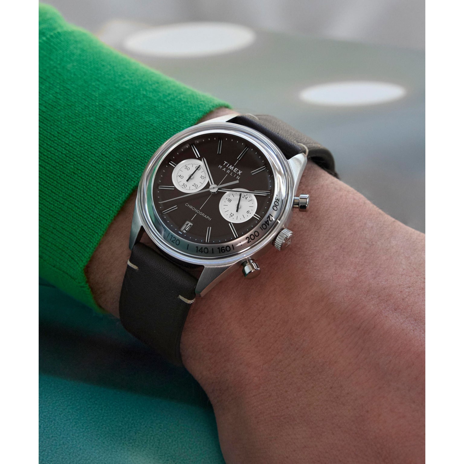 A person wearing a green sleeve shows off a Timex G SS QZ SW JET 40 BROWN DL BWN STP 25 with a domed crystal, black dial, white subdials, and brown leather strap on their left wrist.