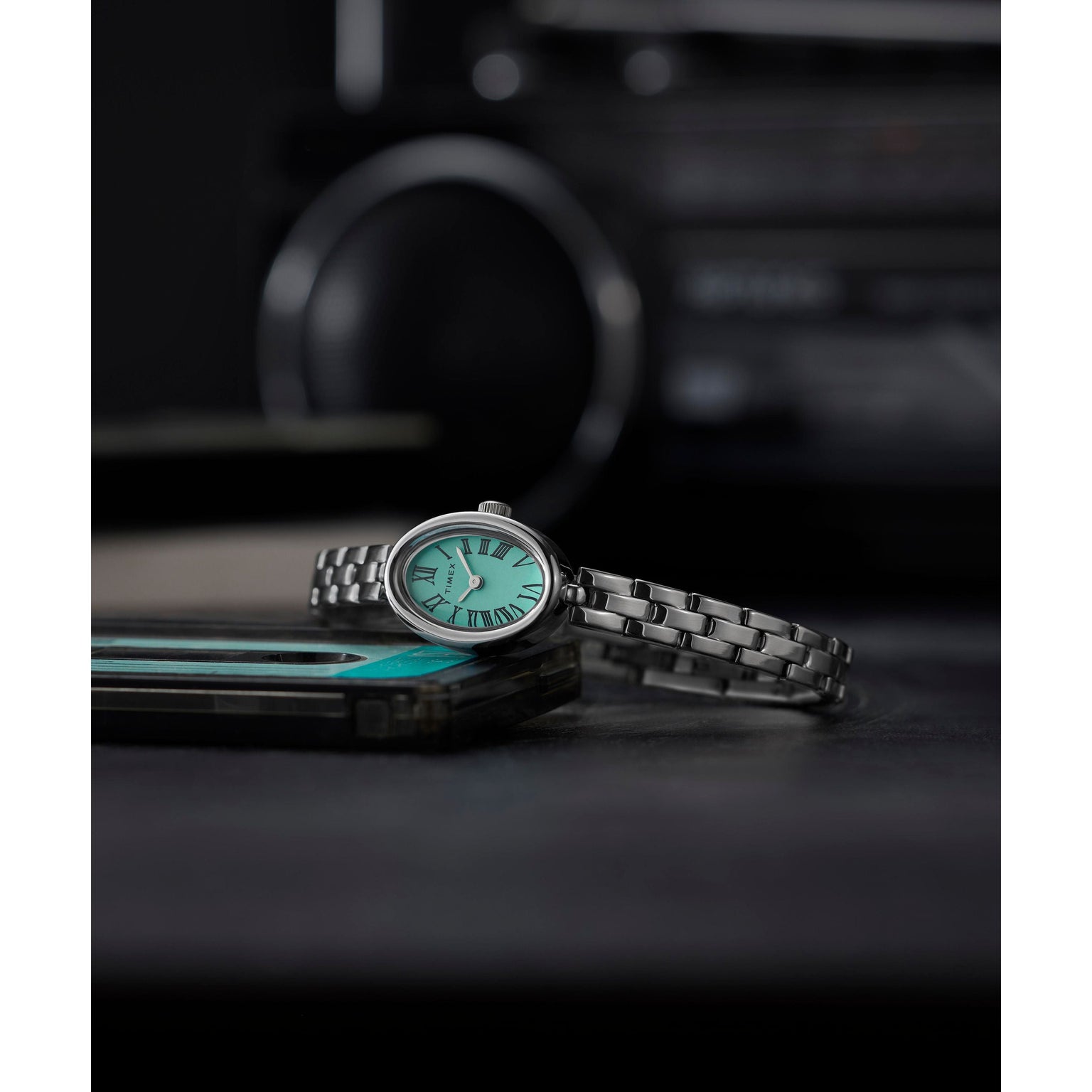 A Timex Cavatina Silver-tone Case and Bracelet watch with an aquamarine dial sits atop a teal and black smartphone, set against a blurred, dark background featuring an electronic device.