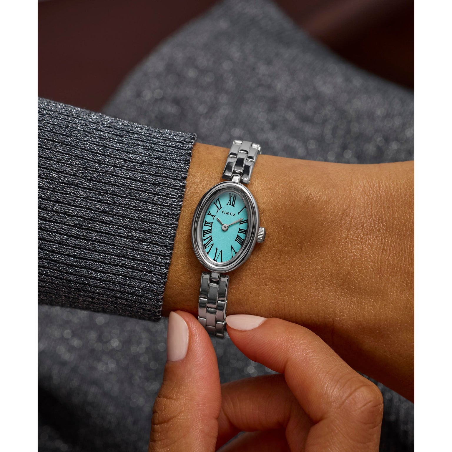 A person wearing a Timex Cavatina Silver-tone Case and Bracelet with Aquamarine Dial, adjusting the watch with their other hand while dressed in a sparkly gray long-sleeve top.