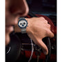 A close-up shows a hand on a car steering wheel, wearing the Timex Q Chronograph with blue accents and a stainless steel bracelet. The watch contrasts with the red dashboard, while the person's face is partly visible.