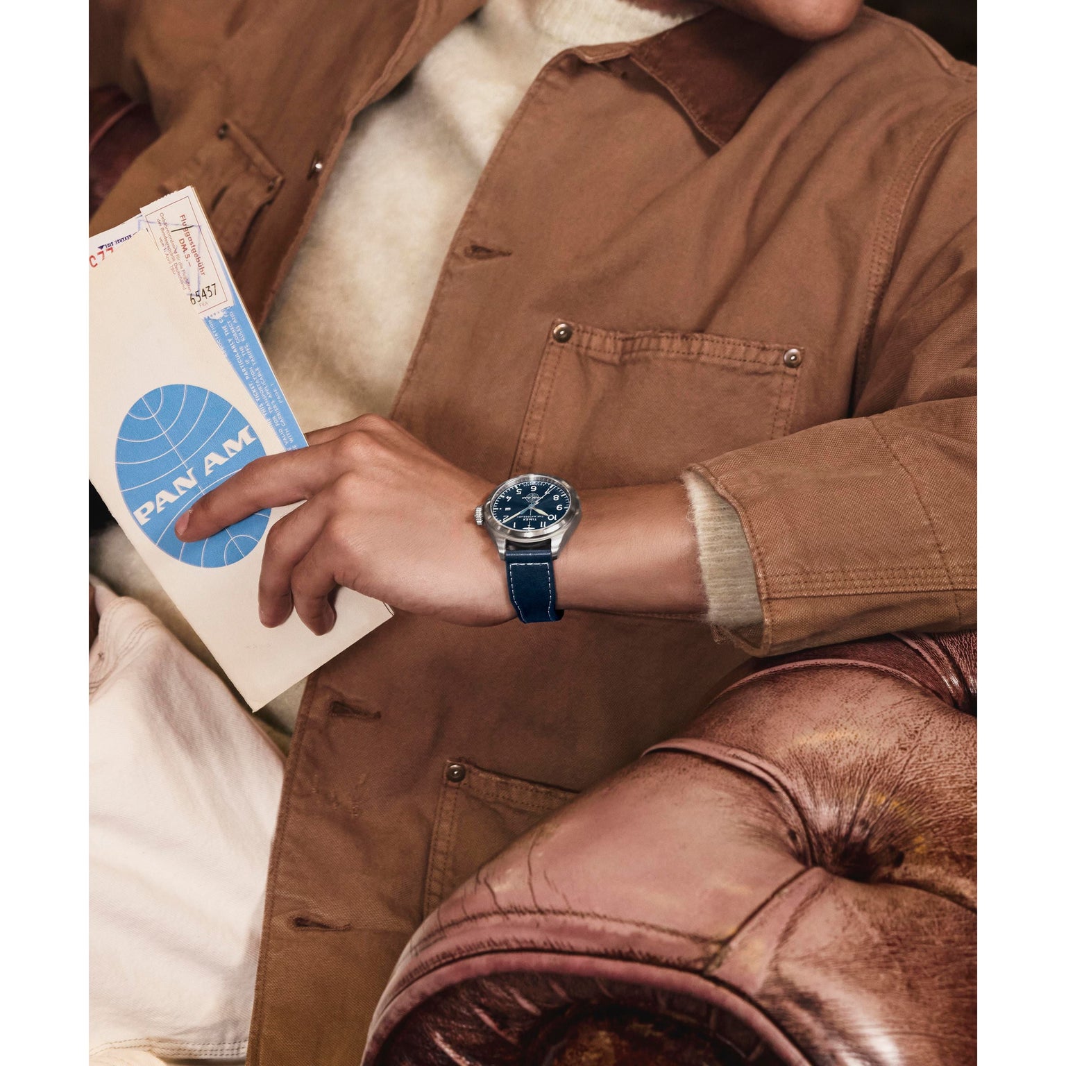 A person in a tan jacket holds a Pan Am envelope, resting their arm on a brown leather chair and wearing the Timex x Pan Am® Waterbury Automatic Ace watch with a blue face and strap by Timex.