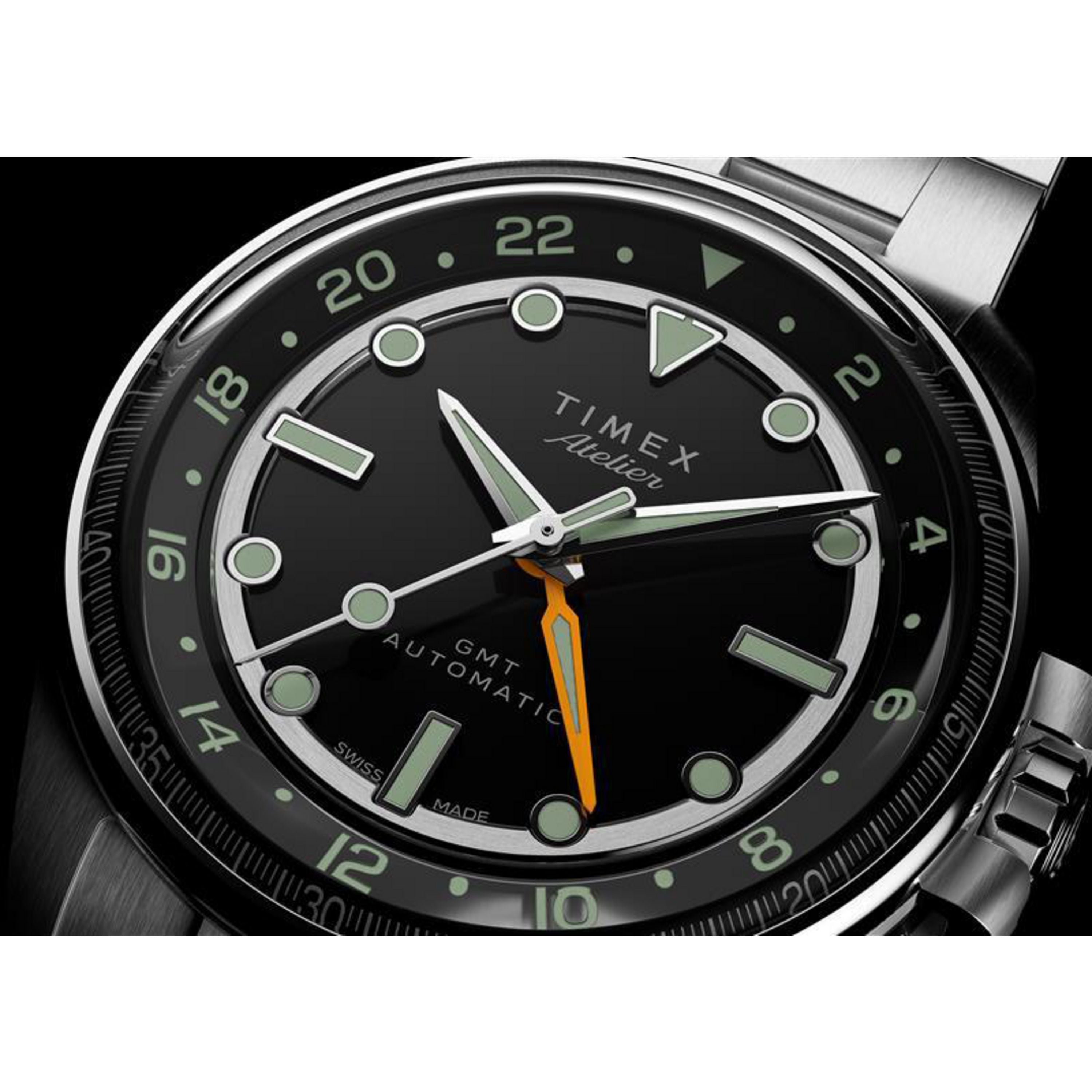 A close-up of the Timex Atelier GMT24 M1a automatic watch, featuring a black dial with silver markers, luminous hands, an orange GMT hand, and "Timex" plus "Automatic" on the face—paired with a stainless steel bracelet.