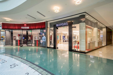 Exterior view of Tudor and Mayors watch stores in a shopping mall, with glass storefronts and displays.