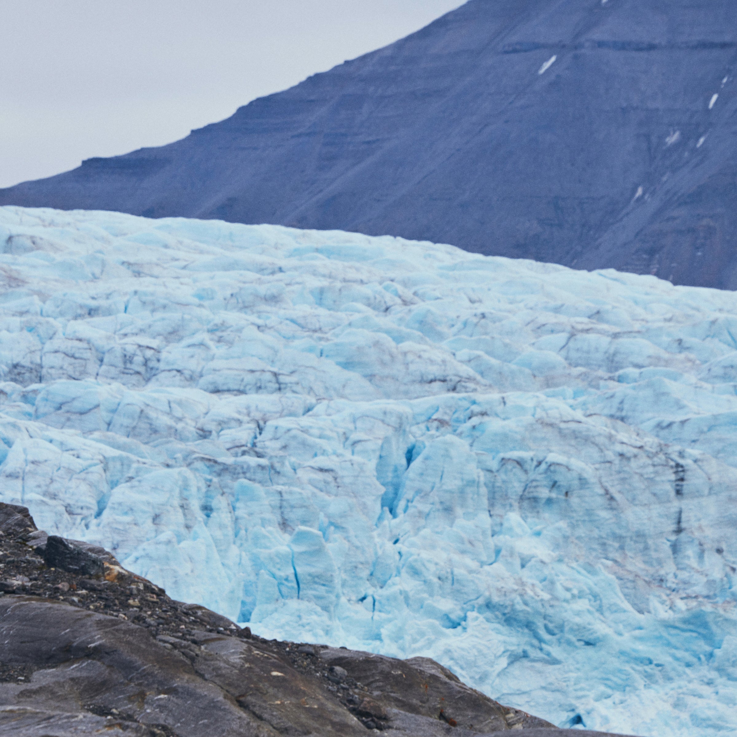 A man stands on a grey rock peak with a glacier in the background, behind the glacier is a mountain with streaks of snow