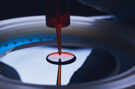 A pipette dispenses red liquid onto a circular glass surface in a laboratory setting.