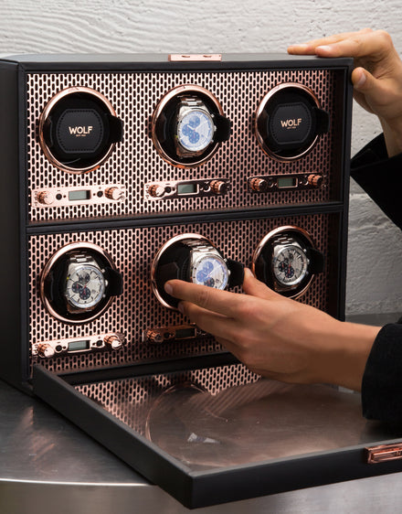 A person places a wristwatch into a luxury watch winder box with multiple slots and a rose gold interior.