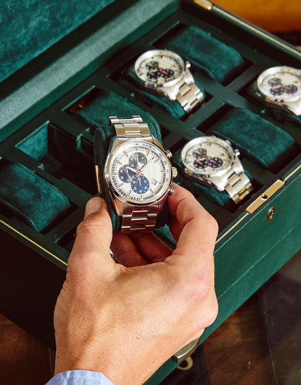 A hand holds a silver luxury watch above a green case containing four similar watches.