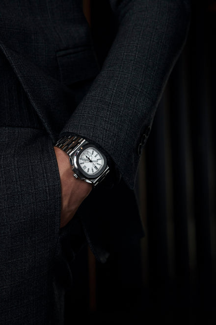 Person in a dark suit with a silver wristwatch, hand partially in pocket, background in shadow.
