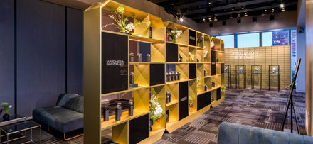 Modern showroom with yellow and black shelving displaying watches, flowers, and decor; city view through windows.