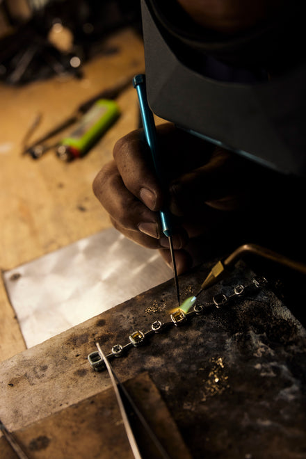 Viewed from above the work space, a person in a welding helmet with a torch works on a pronged jewelry setting
