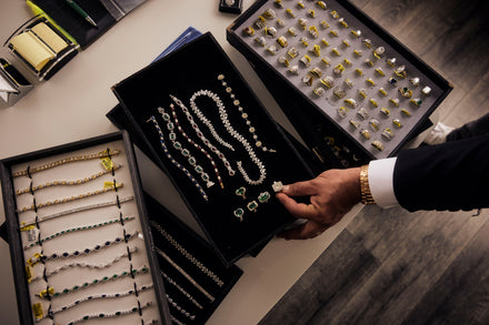 A hand touches a ring in a tray of jewelry next to many trays of rings and bracelets