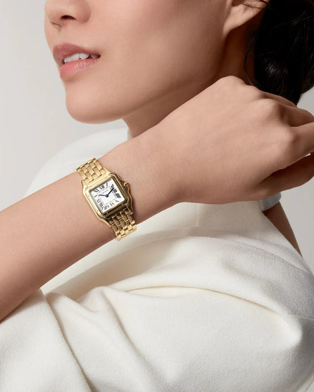 A woman wearing a gold-toned rectangular watch with a white dial and matching gold bracelet, styled with a white top against a light gray studio background.