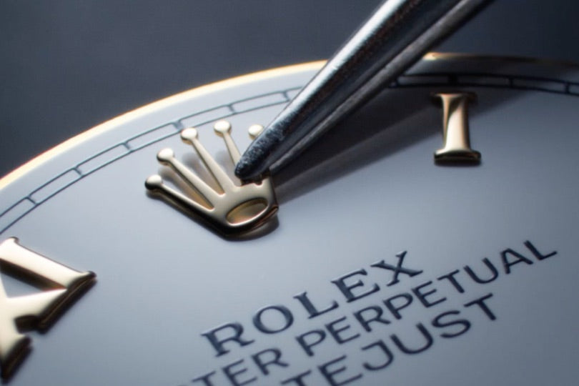 A close-up of tweezers adjusting the gold Rolex crown logo on a watch face.