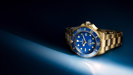 Gold wristwatch with a blue dial and bezel, spotlighted against a dark blue gradient background.