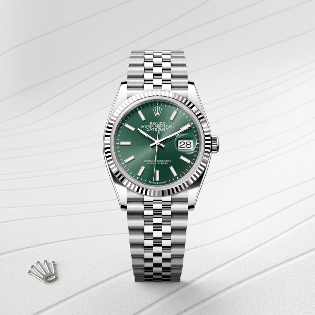 A silver Rolex watch with a green dial and date display on a wavy white surface, with a crown emblem nearby.
