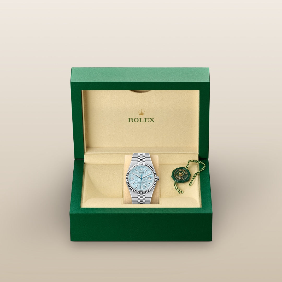 A Rolex watch with a silver band in a green and beige Rolex box, accompanied by a green Rolex tag.