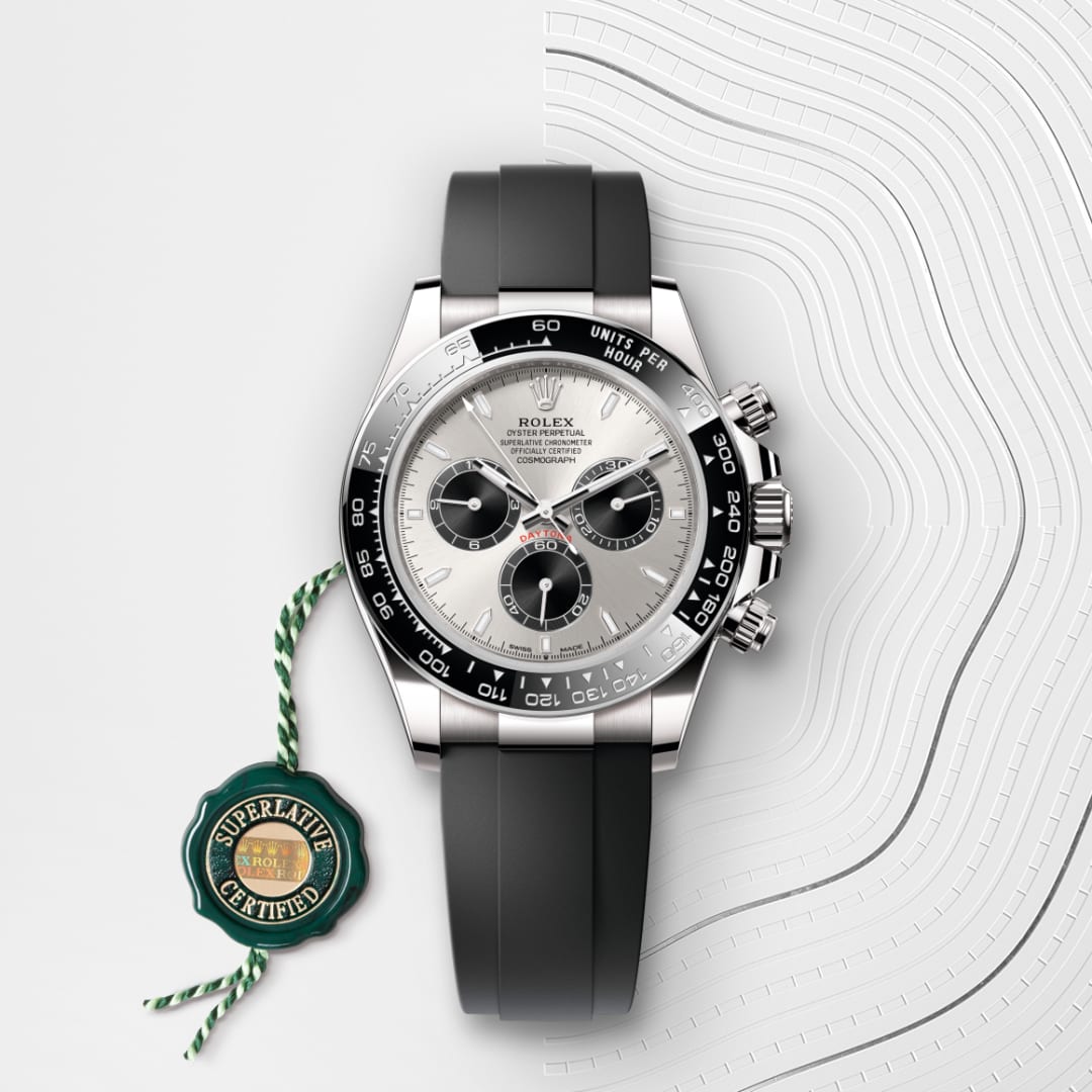 A silver Rolex Daytona watch with a black strap and green Superlative Certified tag on a patterned background.
