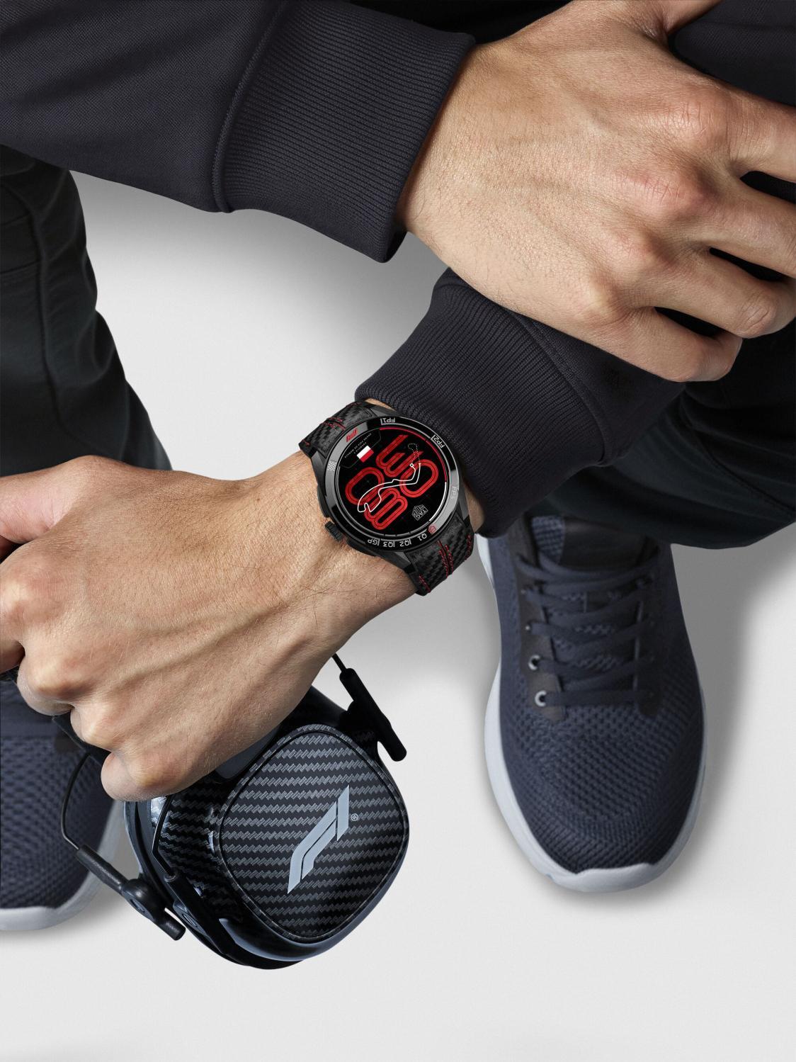 Person wearing a smartwatch with a black case and red display, sitting on a white surface.