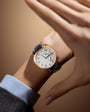 Gold-tone watch with cream dial, blue Arabic numerals, and dark blue leather strap worn on wrist.