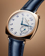 Rose gold cushion-shaped watch with cream dial, blue Arabic numerals, blue hands, and navy leather strap.
