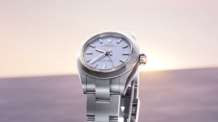 A silver Rolex wristwatch with a light blue face, displayed against a soft, blurred background.