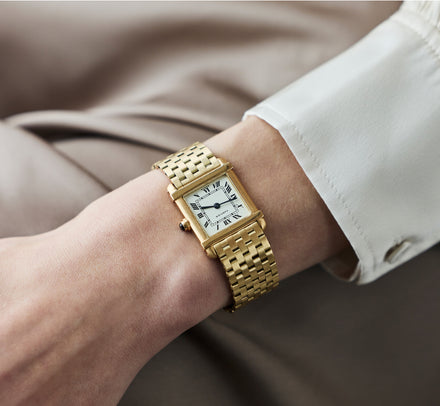 A gold rectangular wristwatch with a white face and Roman numerals on a persons wrist.