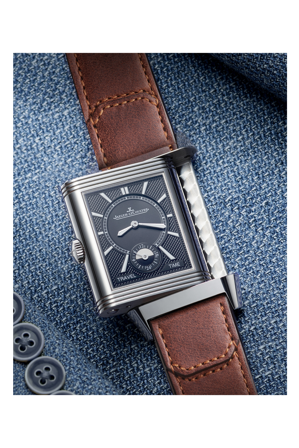 The Jaeger-LeCoultre Reverso Classic Duoface Small Seconds, a rectangular steel luxury watch with a dark patterned dial, silver details, and brown leather strap, sits on blue denim—perfect for tracking two time zones in style.