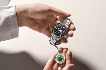 A person holds a silver Rolex wristwatch with a black dial and a green tag in their hands.