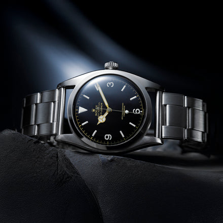 A stainless steel Rolex wristwatch with a black dial, silver strap, and luminous markers, shown on a dark surface.
