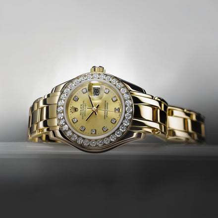A gold luxury wristwatch with a diamond-studded bezel on a reflective surface against a gray background.