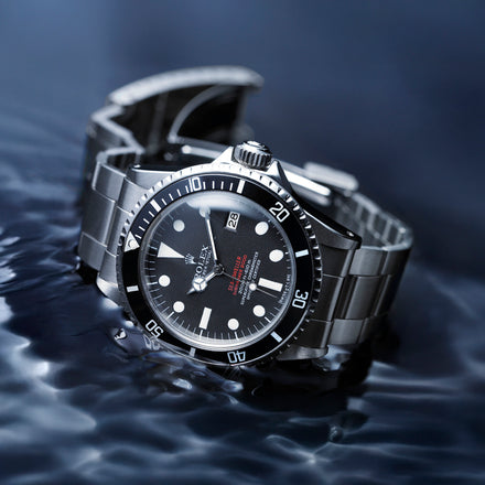 A luxury stainless steel wristwatch with a black dial rests on a rippled water surface.
