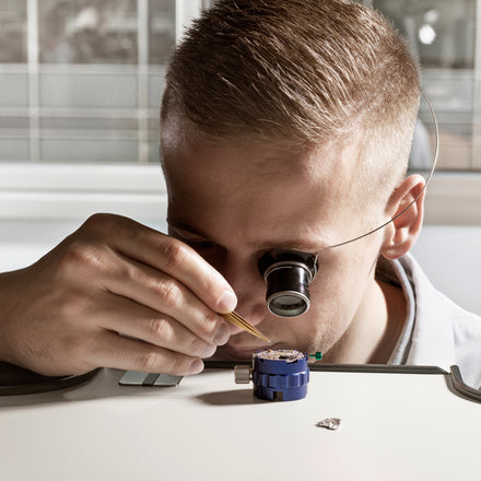 A person uses tweezers and a magnifying loupe to work on a small mechanical device on a desk.