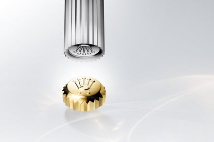 A gold watch crown with the Rolex logo is shown below a silver rod on a light, glossy background.