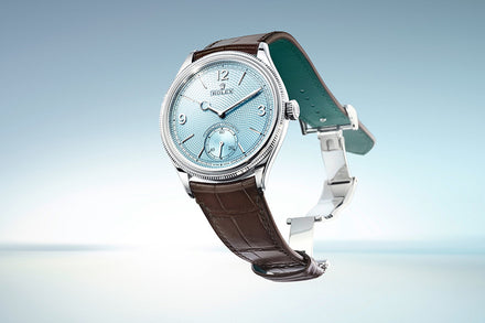 A luxury wristwatch with a light blue dial and brown leather strap against a soft blue background.