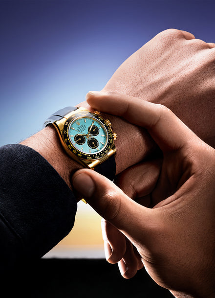 A person adjusts a gold wristwatch with a turquoise face on their left wrist against a twilight sky.