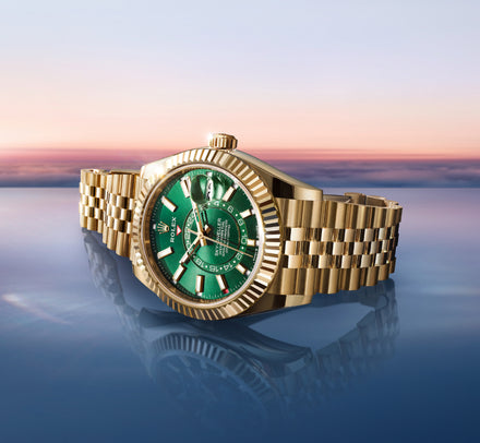 A gold Rolex watch with a green face is displayed against a reflective surface with a sunset sky background.