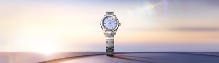 Silver wristwatch with a light blue dial displayed against a soft, sunrise or sunset background.