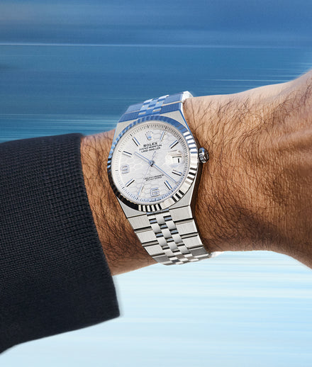 A silver Rolex watch with a white dial on a persons wrist, set against a blurred blue background.
