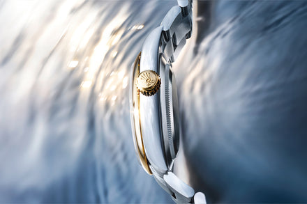 Close-up of a watch with a gold face and white strap on a blurred water background