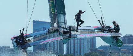 Three sailors racing a T-Mobile-branded sailboat over water, with city buildings in the background.